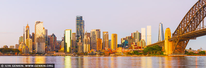 australia stock photography | Sydney City at Dawn, Sydney, New South Wales (NSW), Australia. The photo captures a stunning view of Circular Quay and Sydney City at sunrise, taken from across the harbor. The entire cityscape is bathed in a warm, golden light, which highlights the modern skyline's diverse mix of skyscrapers and historic buildings. The sunlight reflects off the glass facades, casting a soft, amber glow that enhances the architectural details of each structure. Circular Quay, bustling with ferries and boats, is nestled along the water's edge, its iconic wharves shimmering under the sunrise's brilliance. The calm harbor waters mirror the glowing city, creating a breathtaking display of light and color. In the background, the fading sky is a blend of deep oranges, pinks, and purples, adding a sense of drama and serenity to the scene.