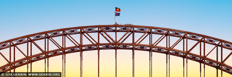 australia stock photography | Panorama of the Sydney Harbour Bridge Silhouette, Sydney, New South Wales, Australia. This panoramic photograph captures the striking arches of the Sydney Harbour Bridge glowing in red as they pierce a serene sky of pale yellow and soft blue. The unique lighting creates an enchanting contrast, with the fiery arches standing out dramatically against the tranquil hues of the sunset. Only the uppermost curves of the bridge are visible, their bold color and elegant form drawing the eye across the expansive composition. This captivating scene highlights the interplay of light, color, and structure, offering a fresh and artistic perspective on Sydney's iconic landmark.