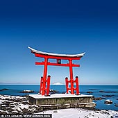  stock photography | Torii Gate of the Toyosaki Konpira Shrine #2, Shosanbetsu, Hokkaido, Japan, Image ID JAPAN-HOKKAIDO-TORII-GATE-0002. A stunning photograph captures the Torii Gate of Toyosaki Konpira Shrine in Shosanbetsu, Hokkaido, Japan. The iconic red gate stands tall against a backdrop of the endless Sea of Japan, its vibrant vermilion hue contrasting beautifully with the azure sky and deep blue waters. The gate's clean, symmetrical lines frame the view beyond, evoking a sense of serenity and connection to the natural world. On the horizon, Mount Rebun on Rebun Island rises majestically, its silhouette adding depth and a sense of distant wonder to the composition. The coastal breeze gently rustles the grasses in the foreground, adding a touch of motion to the scene. The soft light of early morning or late afternoon bathes the gate in a golden glow, enhancing the spiritual and tranquil atmosphere of this sacred location.