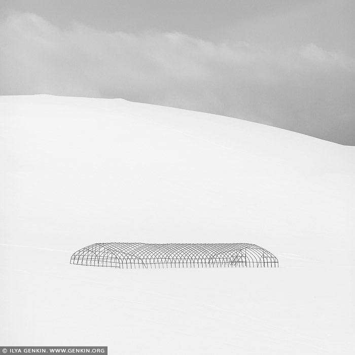 portfolio stock photography | Greenhouse Structure, Study 1, Biei, Hokkaido, Japan. A black-and-white minimalistic photograph captures the stark silhouette of a greenhouse structure standing alone on a snow-covered slope. The clean lines of the greenhouse frame are sharply defined against the pale, featureless expanse of snow, creating a striking contrast between the dark, angular form of the building and the soft, flowing contours of the snowy landscape. The slope of the hill, blanketed in smooth, untouched snow, adds a gentle curve to the composition, while the sky, nearly blending into the whiteness of the ground, remains an empty, bright backdrop. The minimalist nature of the image strips away distractions, leaving only the bold, geometric shape of the greenhouse, isolated and quiet in the vast winter landscape. The silhouette, devoid of detail, is almost abstract, standing in sharp relief against the blank canvas of snow and sky. The scene evokes a sense of stillness and solitude, as if the world outside the frame has disappeared, leaving only the greenhouse and the serene, endless snow. The simplicity of the image highlights both the structureâ€™s fragility and its presence in the vast emptiness, creating a contemplative and peaceful atmosphere.