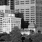 portfolio stock photography | The Astor and Chief Secretary's Building, Sydney, NSW, Australia, Image ID SYDNEY-IN-SQUARE-0025. In this black-and-white photograph, the heritage-listed Chief Secretary's Building, originally and still commonly known as the Colonial Secretary's Building, and The Astor, one of Sydney's most distinguished residential buildings, create a captivating contrast against the modern high-rises of Sydney's evolving skyline. The Chief Secretary's Building stands as a masterpiece of Victorian Free Classical architecture, its richly ornamented facade adorned with detailed pilasters, archways, and a clock tower crowned with a delicate dome. The intricate stonework and classical proportions exude a dignified charm, evoking Sydney's colonial history. Nearby, The Astor — a distinguished residential building in the Interwar Free Classical style — rises with its solid brick and sandstone facade, boasting tall, arched windows and restrained, elegant detailing that emphasise its enduring sophistication. These historic structures are juxtaposed against sleek, contemporary skyscrapers that loom above them, their minimalist glass and steel facades reflecting light in sharp, angular patterns. This striking composition of old and new highlights Sydney's layered architectural identity, where heritage buildings like the Chief Secretary's Building and The Astor retain their presence and dignity amidst the city's continual growth and transformation. The photo captures an architectural dialogue between the historic and the modern, reminding viewers of Sydney's respect for both legacy and innovation.