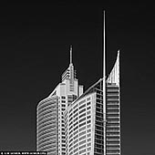 portfolio stock photography | Aurora Place and Chifley Tower, Sydney, NSW, Australia, Image ID SYDNEY-IN-SQUARE-0026. In this black-and-white photograph, Aurora Place and Chifley Tower rise prominently against Sydney's sky, captured in striking detail. Aurora Place, with its curved, sail-like facade, reflects light in varying shades of grey, giving the building a fluid, almost organic presence against the static background. The glass curtain walls of Aurora Place arc gently, with a delicate texture that contrasts its modern, angular neighbouring structures. Beside it stands Chifley Tower, its sleek, stepped design and clean vertical lines embodying the late 20th-century corporate aesthetic. Chifley's smooth concrete and glass surfaces appear slightly darker, grounding it in a timeless, monolithic elegance. The juxtaposition of the two buildings - the organic flow of Aurora Place and the geometric precision of Chifley Tower - illustrates the contrasting architectural philosophies of Sydney's evolving skyline, blending bold modernism with subtle gestures of nature-inspired design.